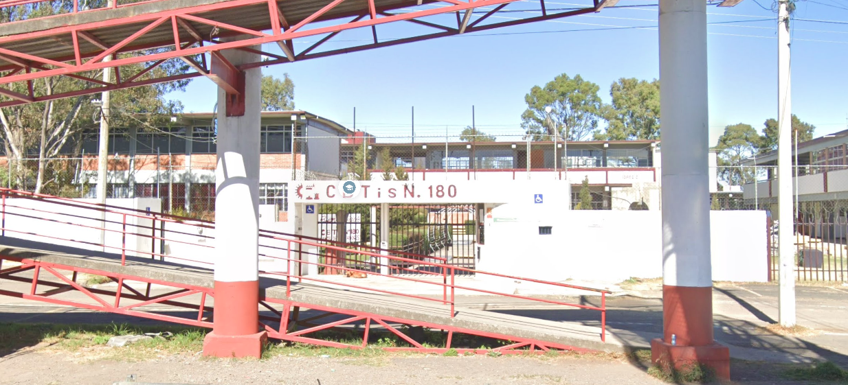 Fotografía en la que se muestra la fachada del plantel, conformada por muros de color blanco que rodean las instalaciones. En medio de los muros se encuentra la reja que da acceso al plantel y sobre ella, en letras rojas, dice Cebetis número 180.